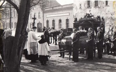 Ksiądz Sobota przyjmuje kondukt pogrzebowy przed kościołem. W tyle widoczne zabudowania ulicy Dworcowej.
